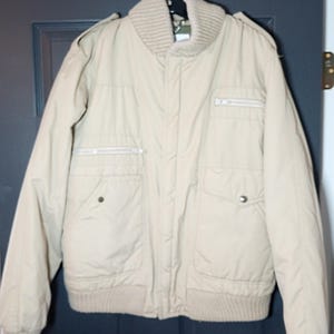 Può includere: Giacca bomber beige con colletto, polsini e orlo a coste. Presenta due tasche anteriori, due tasche sul petto e chiusura con cerniera. La giacca è appesa a una gruccia nera.