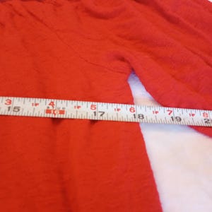Puede incluir: Primer plano de una prenda roja texturizada con mangas largas. La prenda tiene un escote fruncido y un detalle floral decorativo en tonos rosas y morados. Una cinta m&eacute;trica est&aacute; colocada sobre la prenda.