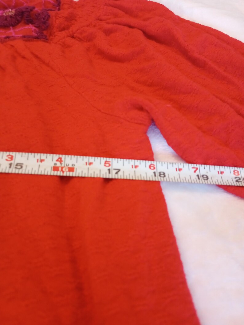 Puede incluir: Primer plano de una prenda roja texturizada con mangas largas. La prenda tiene un escote fruncido y un detalle floral decorativo en tonos rosas y morados. Una cinta m&eacute;trica est&aacute; colocada sobre la prenda.
