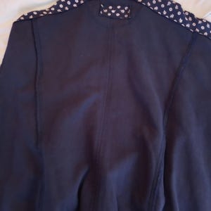 May include: Navy blue jacket with a ribbed collar and patterned shoulder accents. The shoulders feature a white and blue floral design. The jacket has a simple, classic design.