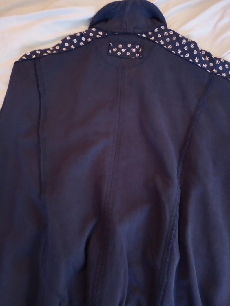 May include: Navy blue jacket with a ribbed collar and patterned shoulder accents. The shoulders feature a white and blue floral design. The jacket has a simple, classic design.