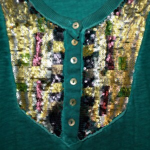 May include: Teal-colored top with a V-neck and a decorative sequined panel. The panel features a mosaic of gold, silver, pink, and black sequins. The top has a button-down placket with four small, round buttons.