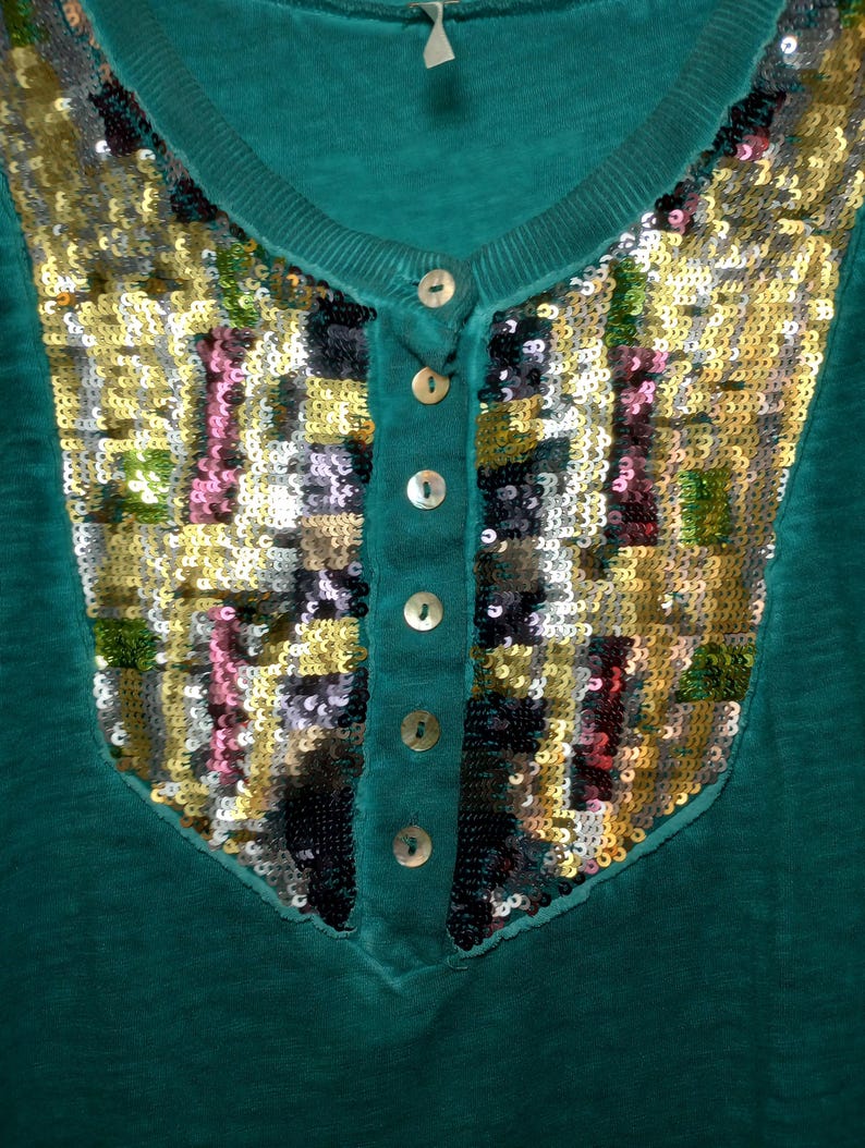 May include: Teal-colored top with a V-neck and a decorative sequined panel. The panel features a mosaic of gold, silver, pink, and black sequins. The top has a button-down placket with four small, round buttons.