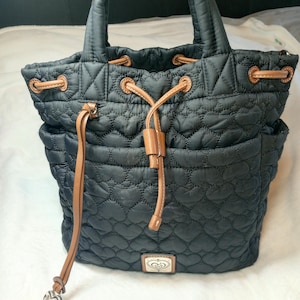 May include: Black quilted tote bag with a heart pattern. Features brown leather accents on the drawstring closure, handles, and side pockets. A silver heart charm hangs from the drawstring. The bag has a small rectangular emblem with a heart design.