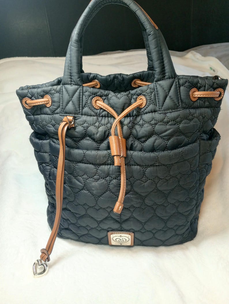 May include: Black quilted tote bag with a heart pattern. Features brown leather accents on the drawstring closure, handles, and side pockets. A silver heart charm hangs from the drawstring. The bag has a small rectangular emblem with a heart design.