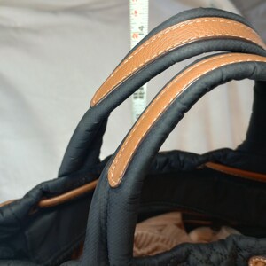 May include: Black quilted tote bag with tan leather handles and trim. The handles are stitched with white thread. The bag has a drawstring closure and a structured design. The bag's height is approximately 15 inches.