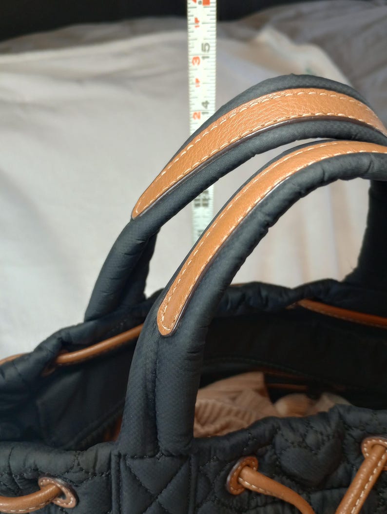 May include: Black quilted tote bag with tan leather handles and trim. The handles are stitched with white thread. The bag has a drawstring closure and a structured design. The bag's height is approximately 15 inches.