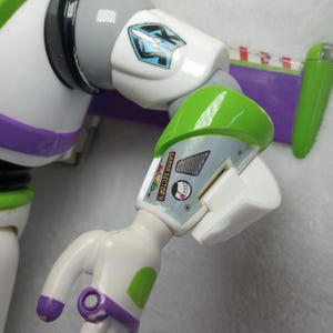 May include: A plastic action figure arm featuring a white, green, and purple color scheme. The upper arm displays a blue and white rocket emblem, while the forearm includes a graphic label with the text GAMMA SECTOR 4 and a circular gauge.