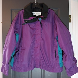 Könnte beinhalten: Eine lila Columbia Jacke mit türkisfarbenen Seitenteilen und einem schwarzen Kragen. Die Jacke hat einen Reißverschluss vorne, zwei Vordertaschen und das Columbia-Logo auf der Brust. Die Jacke hängt an einem schwarzen Kleiderbügel.