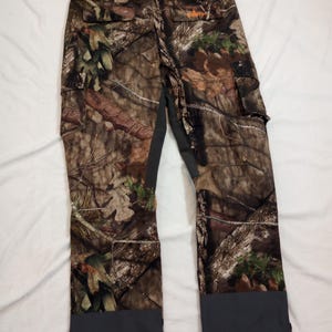 Puede incluir: Pantalones de caza de camuflaje con un patr&oacute;n marr&oacute;n, verde y beige. Los pantalones tienen bolsillos de carga y detalles grises en la cintura y los tobillos. Un logotipo naranja es visible en la parte trasera.