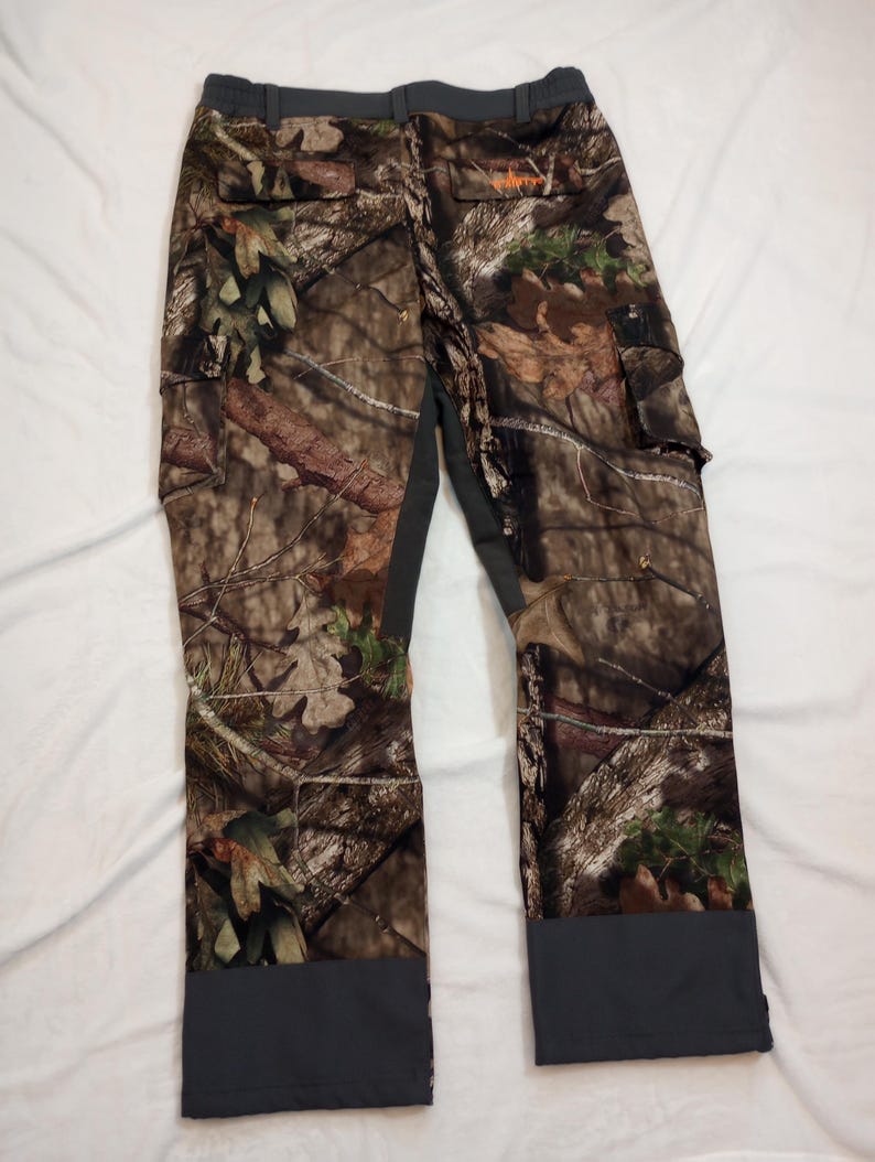 Puede incluir: Pantalones de caza de camuflaje con un patr&oacute;n marr&oacute;n, verde y beige. Los pantalones tienen bolsillos de carga y detalles grises en la cintura y los tobillos. Un logotipo naranja es visible en la parte trasera.