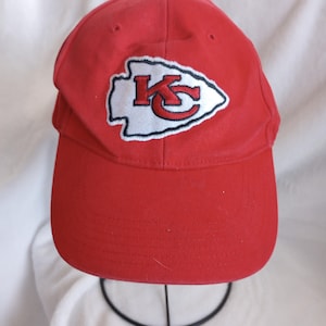 May include: A red baseball cap featuring the Kansas City Chiefs logo. The logo is an arrowhead shape with the letters "KC" in red, outlined in white and black. The cap is made of a durable fabric.