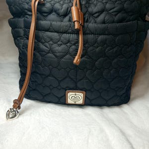 May include: Black quilted drawstring bag with a heart pattern. Features brown leather-like drawstring and accents. A silver heart charm hangs from the side. The bag has a small rectangular patch with a logo.