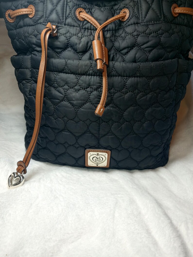 May include: Black quilted drawstring bag with a heart pattern. Features brown leather-like drawstring and accents. A silver heart charm hangs from the side. The bag has a small rectangular patch with a logo.