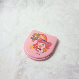 May include: A pink, vintage compact mirror featuring a cartoon girl with pink hair, sunglasses, and a flower. A rainbow and a cartoon dog are also depicted. The compact is closed and has rounded edges. The text "©M.I. 1982" is printed on the compact.