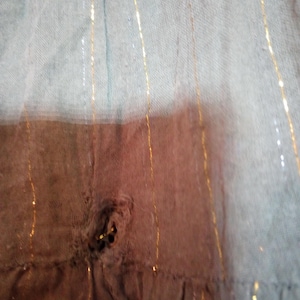K&ouml;nnte beinhalten: Nahaufnahme eines verwaschenen, hellblauen Stoffes mit vertikalen goldenen Metallic-Streifen. Der Stoff hat ein Loch in der Mitte, das eine dunklere braune Schicht darunter freilegt. Der untere Rand hat eine R&uuml;sche.