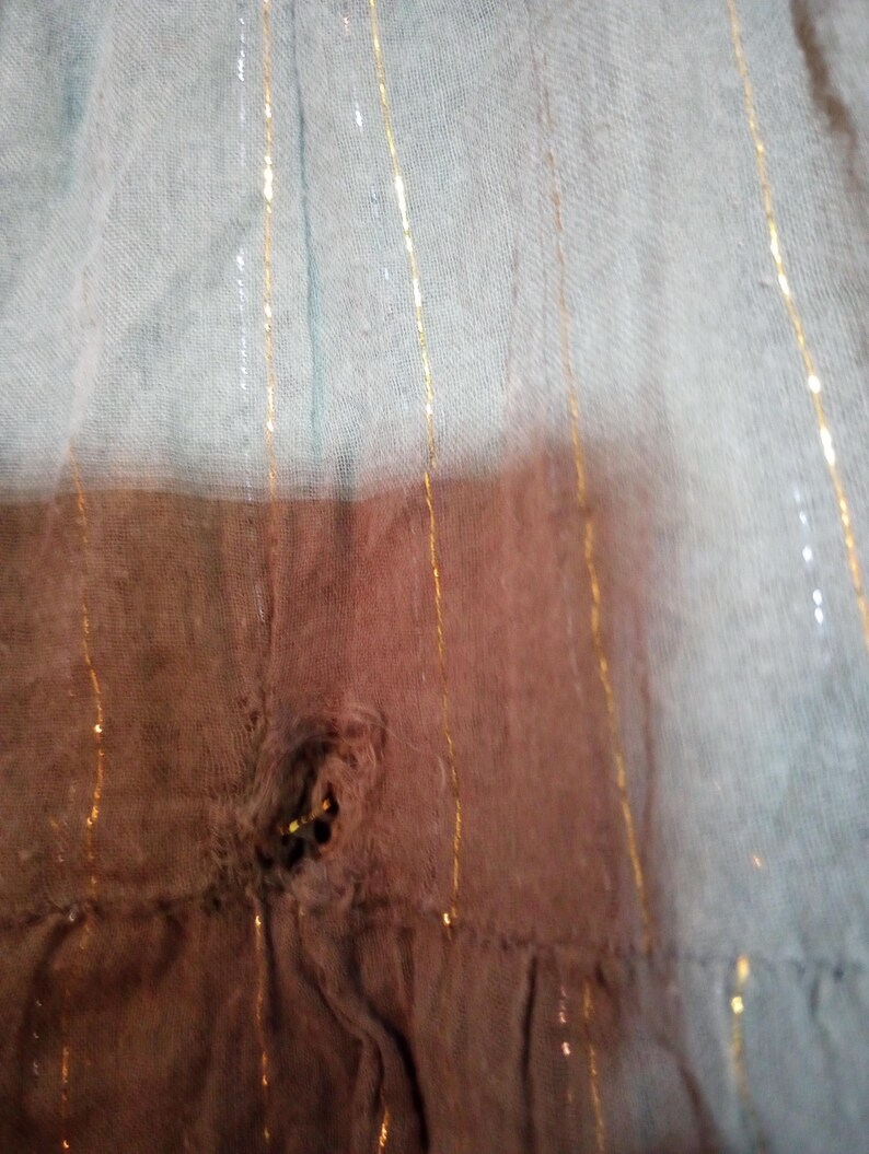 K&ouml;nnte beinhalten: Nahaufnahme eines verwaschenen, hellblauen Stoffes mit vertikalen goldenen Metallic-Streifen. Der Stoff hat ein Loch in der Mitte, das eine dunklere braune Schicht darunter freilegt. Der untere Rand hat eine R&uuml;sche.