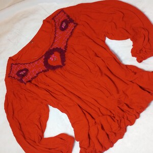 Puede incluir: Una blusa roja vibrante con mangas largas y dobladillo con volantes. El escote presenta intrincados detalles bordados en tonos burdeos y rosa, agregando un toque decorativo a la prenda. La blusa est&aacute; hecha de una tela suave y fluida.