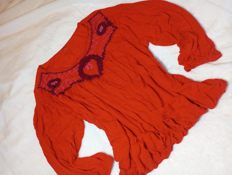 Puede incluir: Una blusa roja vibrante con mangas largas y dobladillo con volantes. El escote presenta intrincados detalles bordados en tonos burdeos y rosa, agregando un toque decorativo a la prenda. La blusa est&aacute; hecha de una tela suave y fluida.