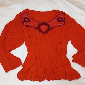 Puede incluir: Una blusa roja de manga larga con cuello redondo y bajo con volantes. El canes&uacute; est&aacute; decorado con detalles bordados en tonos rosa y burdeos. Las mangas est&aacute;n recogidas en los pu&ntilde;os.