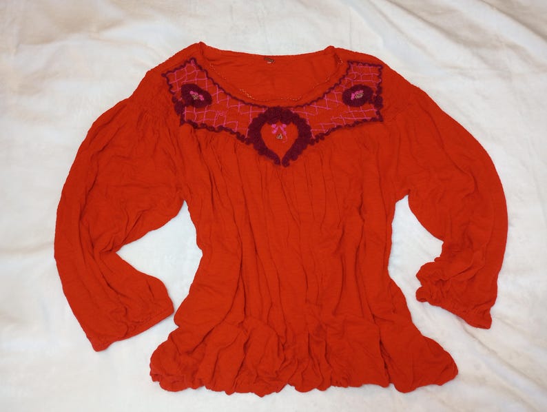 Puede incluir: Una blusa roja de manga larga con cuello redondo y bajo con volantes. El canes&uacute; est&aacute; decorado con detalles bordados en tonos rosa y burdeos. Las mangas est&aacute;n recogidas en los pu&ntilde;os.