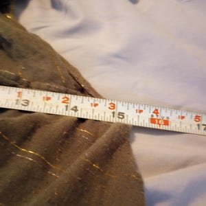 K&ouml;nnte beinhalten: Nahaufnahme eines olivgr&uuml;nen Stoffes mit goldenen Fadenakzenten. Ein Ma&szlig;band zeigt eine L&auml;nge von 33 bis 43 cm. Der Stoff scheint Teil eines Kleidungsst&uuml;cks zu sein.