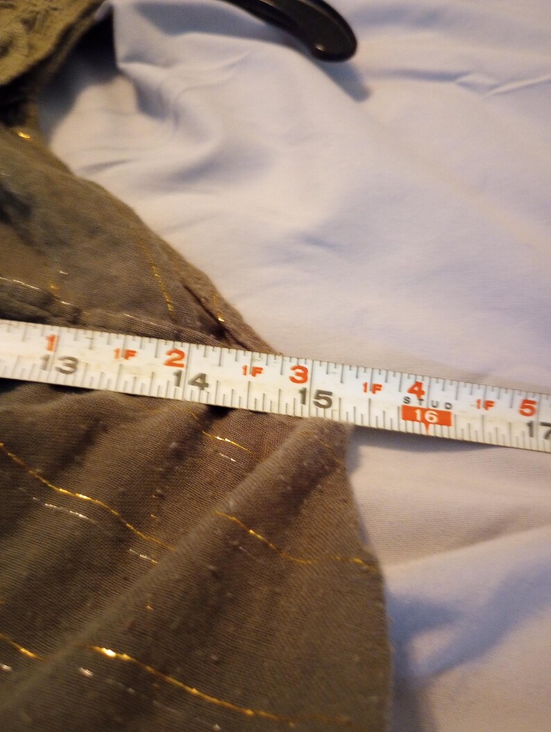 K&ouml;nnte beinhalten: Nahaufnahme eines olivgr&uuml;nen Stoffes mit goldenen Fadenakzenten. Ein Ma&szlig;band zeigt eine L&auml;nge von 33 bis 43 cm. Der Stoff scheint Teil eines Kleidungsst&uuml;cks zu sein.
