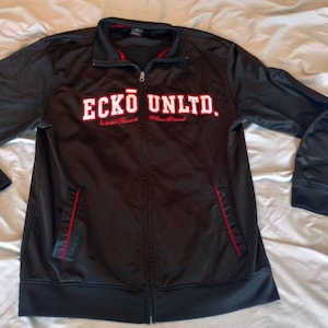 May include: Black zip-up jacket with the text "ECKO UNLTD." in white block letters. The jacket has red accents on the pockets and collar. The sleeves are long and the jacket is made of a smooth material.