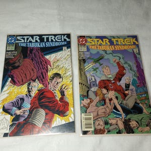 Puede incluir: Se muestran dos cómics de Star Trek, "El Síndrome Tabukan". Uno presenta una figura vestida de rojo, el otro a un hombre de rojo con otros en apuros. Los cómics tienen ilustraciones coloridas y las portadas están en fundas protectoras.