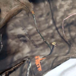 Puede incluir: Primer plano de tela de camuflaje con el logotipo de Mossy Oak y la palabra HABIT en naranja. La tela presenta un patr&oacute;n marr&oacute;n y verde con detalles de ramas de &aacute;rboles, adecuada para actividades al aire libre.