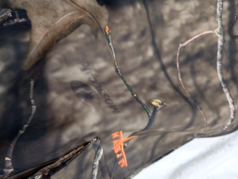 Puede incluir: Primer plano de tela de camuflaje con el logotipo de Mossy Oak y la palabra HABIT en naranja. La tela presenta un patr&oacute;n marr&oacute;n y verde con detalles de ramas de &aacute;rboles, adecuada para actividades al aire libre.