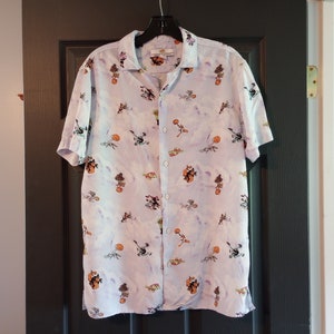 Puede incluir: Una camisa de botones azul claro con un estampado tie-dye blanco y azul claro. La camisa presenta un estampado de personajes de dibujos animados con una variedad de personajes en diferentes poses.