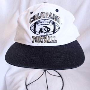 Puede incluir: Gorra de béisbol blanca y negra con la inscripción "COLORADO FOOTBALL" bordada en oro y negro. La gorra tiene una visera negra y un gráfico de fútbol americano. El sombrero se exhibe en un soporte de metal negro.