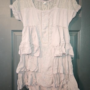 May include: A light mauve-colored dress with a lace yoke and short sleeves. The dress features embroidered detailing down the front and layered ruffles. The dress is hanging on a black hanger against a dark green door.