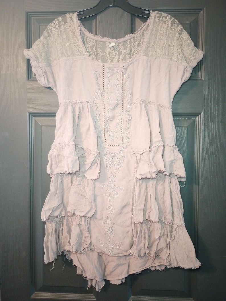 May include: A light mauve-colored dress with a lace yoke and short sleeves. The dress features embroidered detailing down the front and layered ruffles. The dress is hanging on a black hanger against a dark green door.