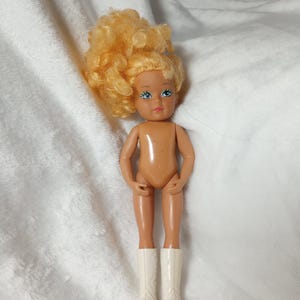 May include: A small doll with blonde, curly hair and blue eyes. The doll has a tan body and is wearing white boots. The doll is posed standing up. The doll is a vintage toy.