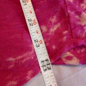 May include: Close-up of a vibrant fuchsia fabric with a textured pattern and light beige accents. A measuring tape shows a length of approximately 53 to 64 cm. The fabric appears to be a soft material.