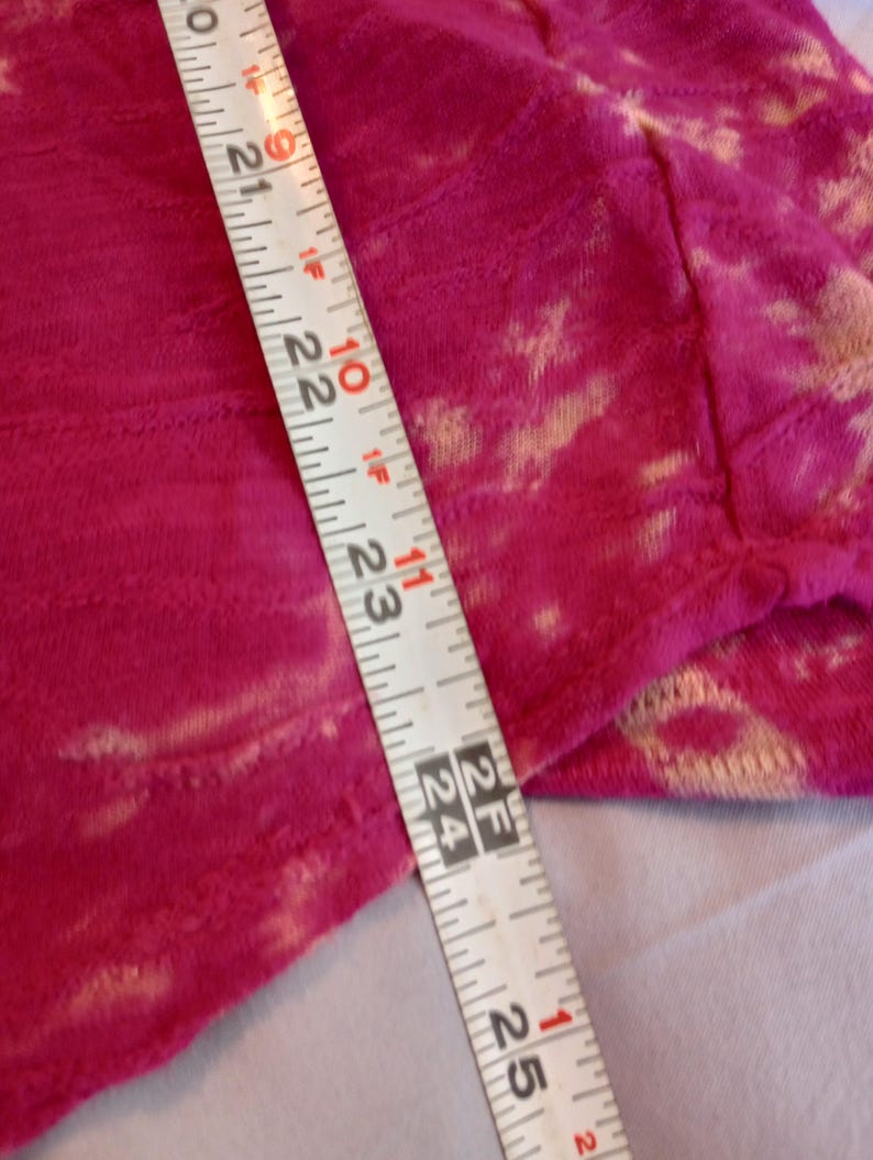 May include: Close-up of a vibrant fuchsia fabric with a textured pattern and light beige accents. A measuring tape shows a length of approximately 53 to 64 cm. The fabric appears to be a soft material.
