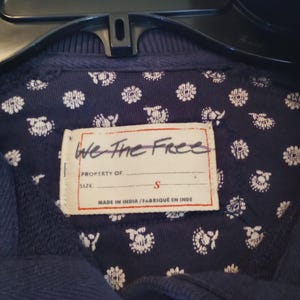 May include: Close-up of a navy blue garment with a floral patterned lining. The label reads "We The Free" and indicates a size "S". The garment is hanging on a black hanger.