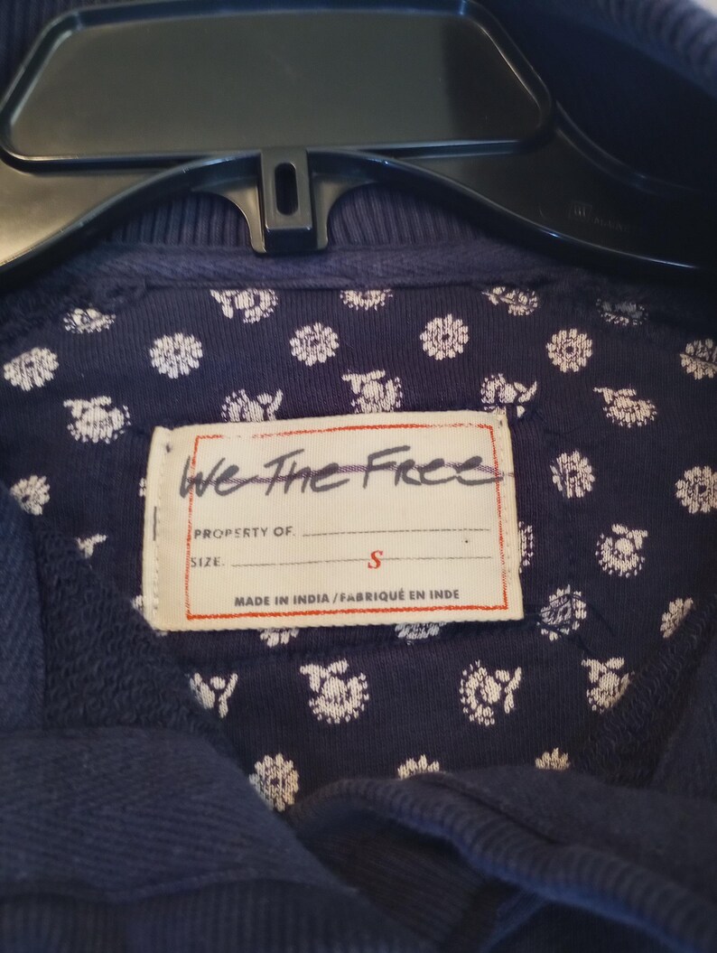 May include: Close-up of a navy blue garment with a floral patterned lining. The label reads "We The Free" and indicates a size "S". The garment is hanging on a black hanger.