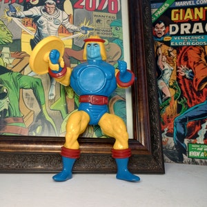 Puede incluir: Una figura de acción de plástico azul y amarillo de un superhéroe sosteniendo un platillo. La figura tiene un cinturón rojo y botas rojas y amarillas. La figura está de pie frente a una portada de cómic con el texto "Manhunter 2070".
