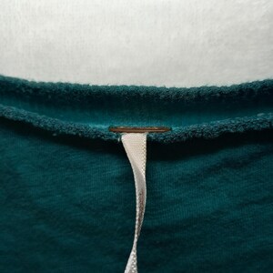 May include: Close-up of a teal-colored garment with a textured surface. A small, round, copper-colored detail is visible near the neckline. A white ribbon-like tag hangs down from the garment.