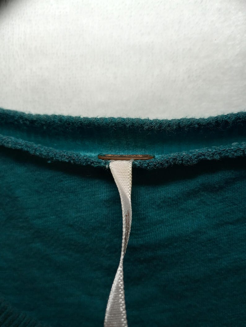 May include: Close-up of a teal-colored garment with a textured surface. A small, round, copper-colored detail is visible near the neckline. A white ribbon-like tag hangs down from the garment.