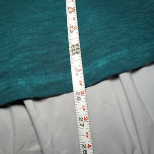 May include: A teal-colored fabric is measured with a white measuring tape. The tape shows measurements from 20 to 29 inches. The fabric is textured and appears to be a piece of clothing.