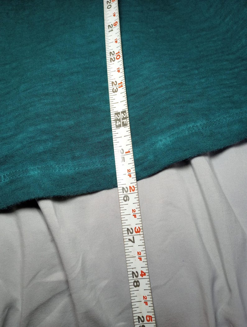 May include: A teal-colored fabric is measured with a white measuring tape. The tape shows measurements from 20 to 29 inches. The fabric is textured and appears to be a piece of clothing.