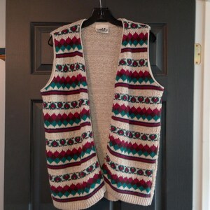 May include: A cream-colored knitted vest with a geometric pattern in shades of teal, burgundy, and white. The vest has a V-neck and a loose fit.