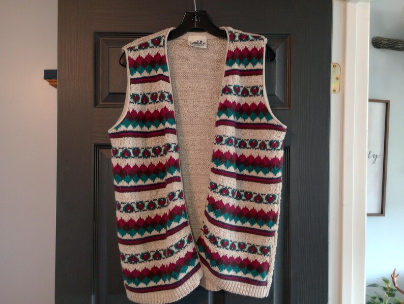 May include: A cream-colored knitted vest with a geometric pattern in shades of teal, burgundy, and white. The vest has a V-neck and a loose fit.