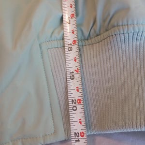 Może przedstawiać: Jasnoniebieska kurtka ze ściągaczem. Kurtka jest mierzona linijką, pokazując długość około 53 cm.