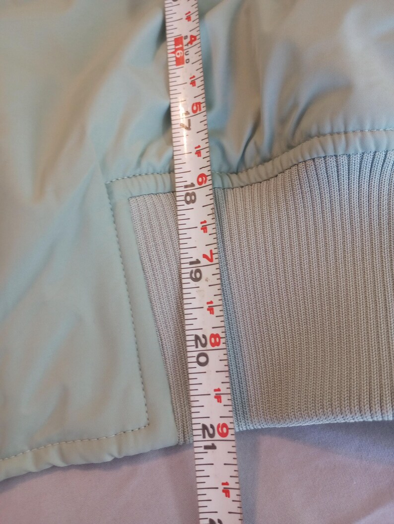 Może przedstawiać: Jasnoniebieska kurtka ze ściągaczem. Kurtka jest mierzona linijką, pokazując długość około 53 cm.