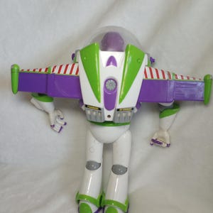 May include: A plastic action figure of a space ranger character with a purple head, white body, and green accents. The figure features extended purple wings with red and white striped detailing, a clear dome helmet, and labels reading DANGER and JET EXHAUST.